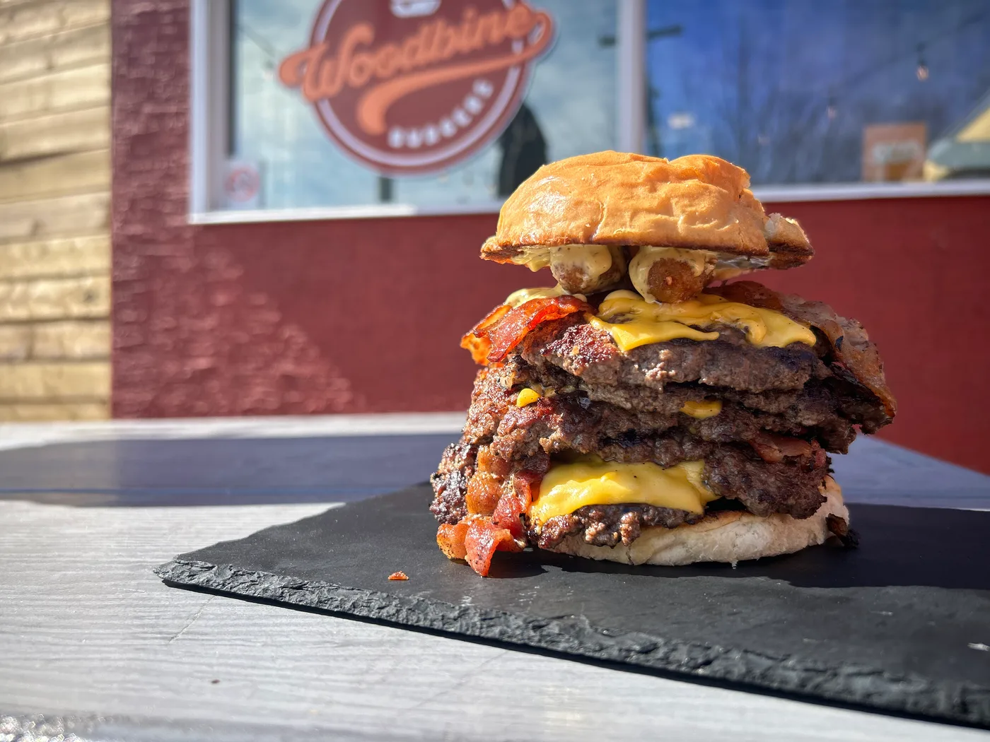 A triple-patty bacon smash burger on slate in front of the Woodbine window