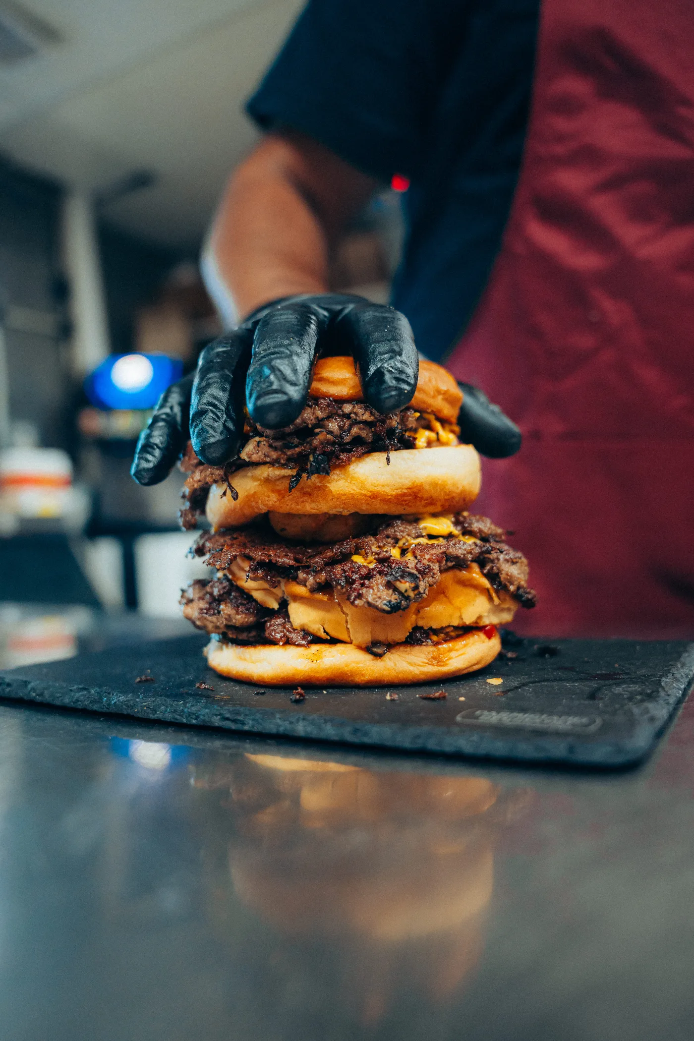A gloved hand pressing down on a triple-patty smash burger on slate